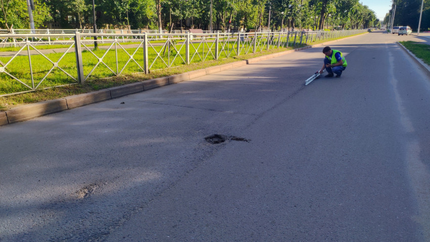 На пустой полосе видна большая выбоина, а инспектор присел с рейкой у ограждения