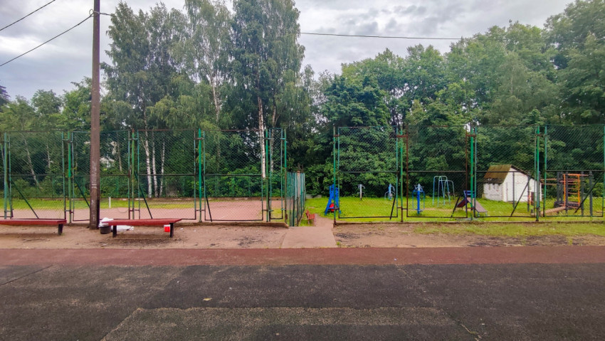 Вход на огороженную спортплощадку с лавками и тренажёрами на фоне деревьев
