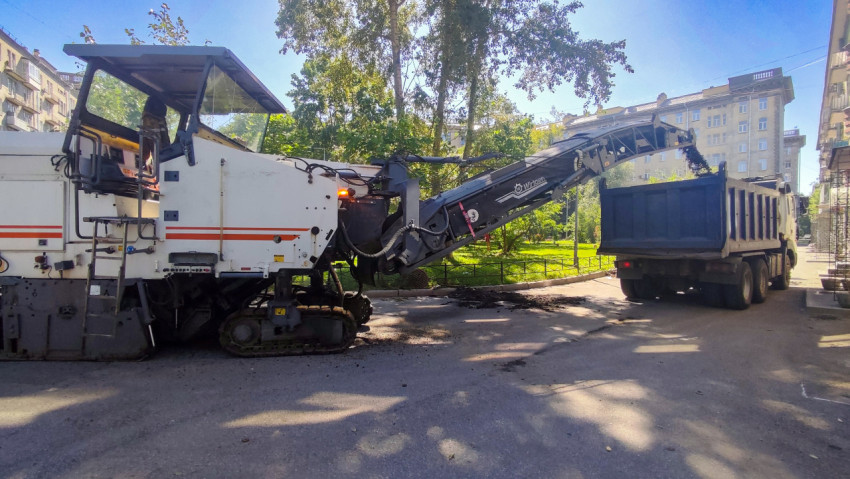 Дорожная фреза Wirtgen загружает снятый асфальт в самосвал на фоне зелёного двора и домов