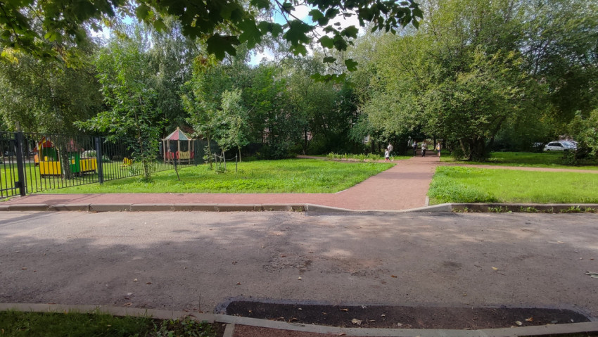 За дорогой видно красную дорожку и детскую площадку за высоким забором среди деревьев