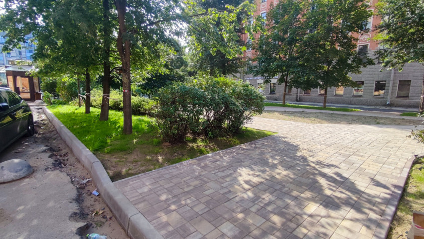 Поворот тротуара из плитки с бордюрами, кустарниками и деревьями возле парковки